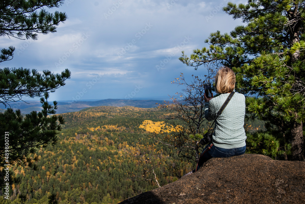 A girl sits on a mountain ledge, admiring the autumn foliage on the neighboring mountains. Beautiful landscape, tourism