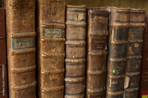Dusty, worn and faded leather-bound Jewish holy books stacked upright on a library shelf.