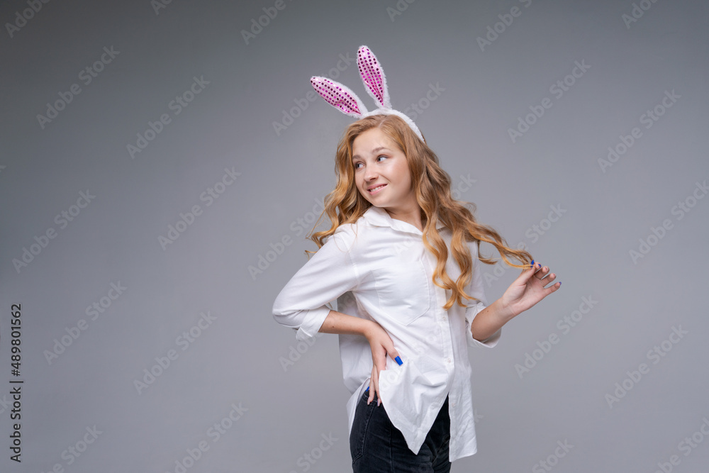 Young beautiful woman in cute easter bunny ears makes a normal gesture, plays with her hand with long hair, in a white shirt on a gray background smiles happily. Caucasian girl in the studio