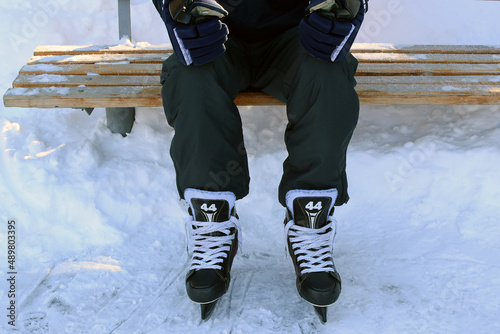  Man in hockey skates and hockey gloves sits on a bench outdoors. Skate lacing.