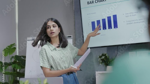 Confident Indian woman manager coach speaker presenting marketing project on flip chart training business people at seminar event give TV chart room presentation during corporate workshop concept.