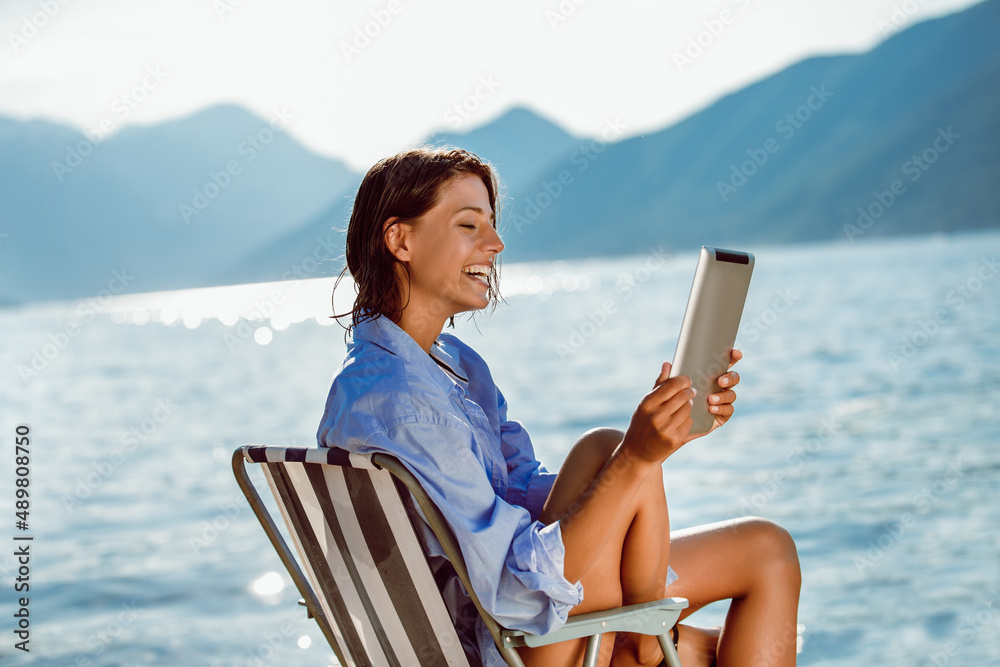 Smiling woman sitting on deck chair by the sea using tablet on a sunny day Smiling woman sitting on deck chair by the sea using tablet on a sunny day