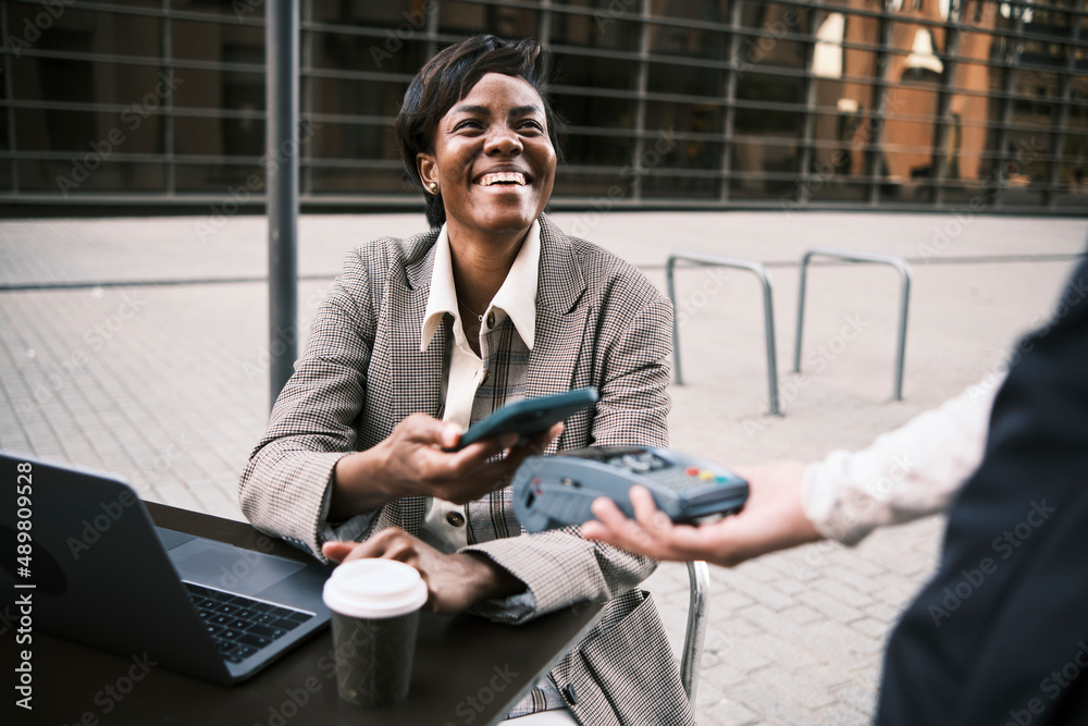 Happy businesswoman making contactless payment through smart phone to waitress at sidewalk cafe