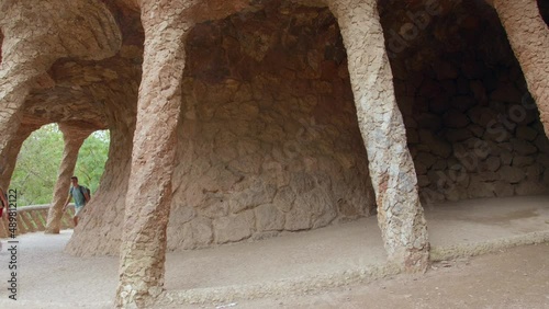 Couple Tourists Walking Under Colonnaded Pathway In Park Guell, Barcelona, Catalonia, Spain - wide shot