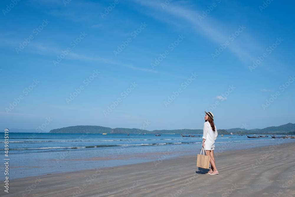 Portrait image of a beautiful young asian woman with hat and bag strolling on the beach with the sea and blue sky background Portrait image of a beautiful young asian woman with hat and bag strolling on the beach with the sea and blue sky background