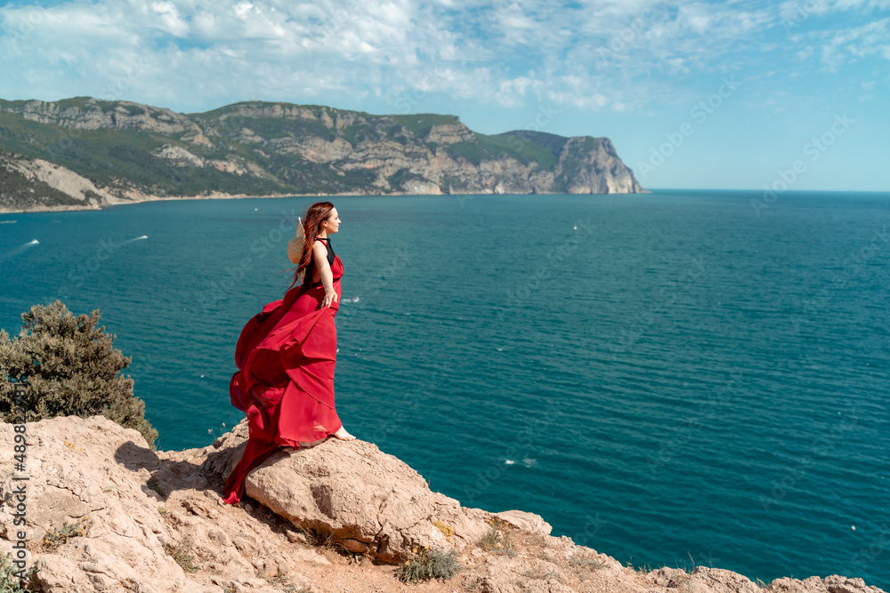 A woman, a side view in a red flying dress fluttering in the wind, a girl in a fluttering dress on the background of the sea. A straw hat hangs at the back of the neck. The concept of a vacation at
