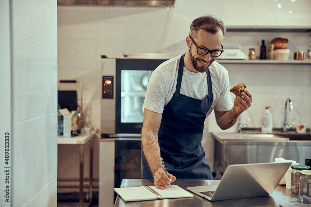 Cheerful man holding pastry and writing in notebook