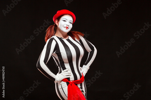 Portrait of female mime artist performing, isolated on black background. Woman stands with her hands on her sides, posing in striped dress smiling