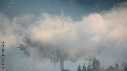 Elevated view of fog moving around small island on lake Bled, Slovenia. Aerial, telephoto perspective of famous Assumption of Maria Church tower. Fog or mist on the surface. Static shot, real time