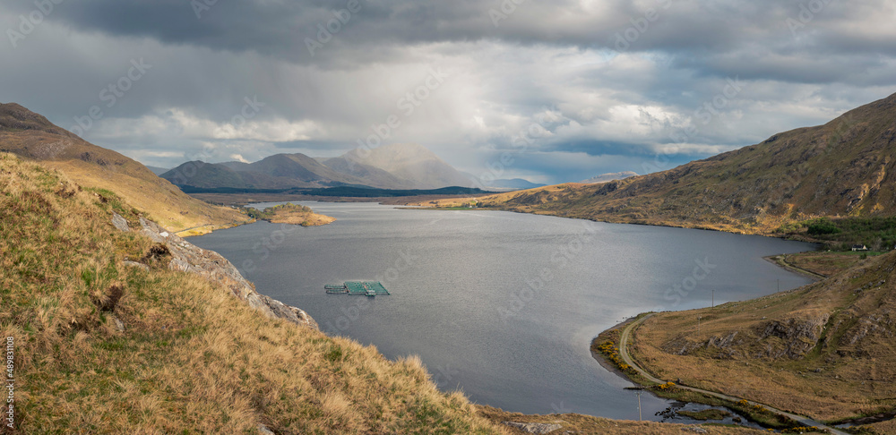 Panorama image of Lough Fee in Connemara, county Galway, Ireland. Beautiful lake surrounded by mountains, Cloudy sky. Calm and ethereal atmosphere. Fish farm. Rain over mountains in the background.