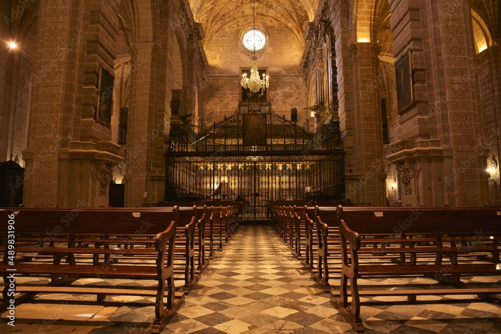 Impressive view through symmetrical wooden church benches and black white tiled checkered floor leading up to the barred choir area of Iglesia Mayor Prioral at Puerto de Santa María, Spain