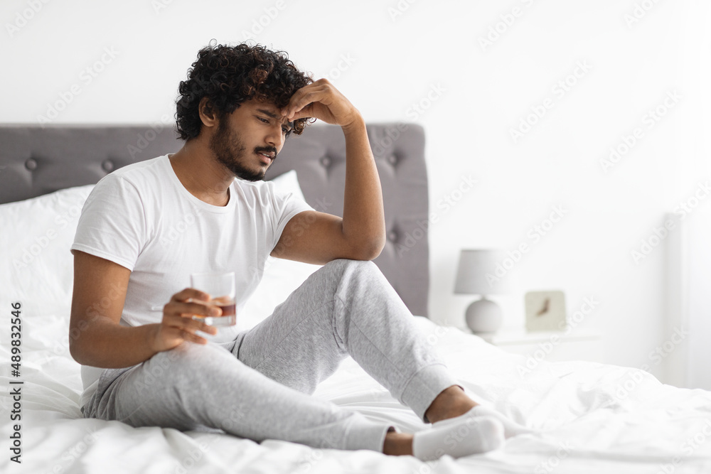 Frustrated indian guy drinking alcohol, sitting on bed, copy space Frustrated indian guy drinking alcohol, sitting on bed, copy space