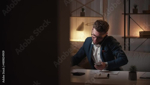 Business tasks. Busy man. Financial accounting. Elegant rich guy counting cash money talking mobile phone sitting desk in dark light room interior. photo