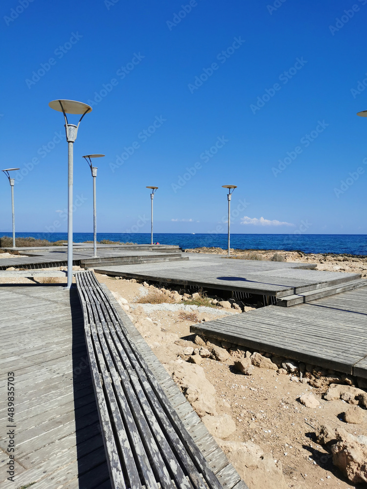 A boardwalk with a wooden bench and lanterns along the rocky coast of the Mediterranean Sea.