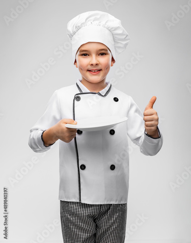 cooking, culinary and profession concept - happy smiling little boy in chef's toque and jacket holding empty plate showing thumbs up over grey background