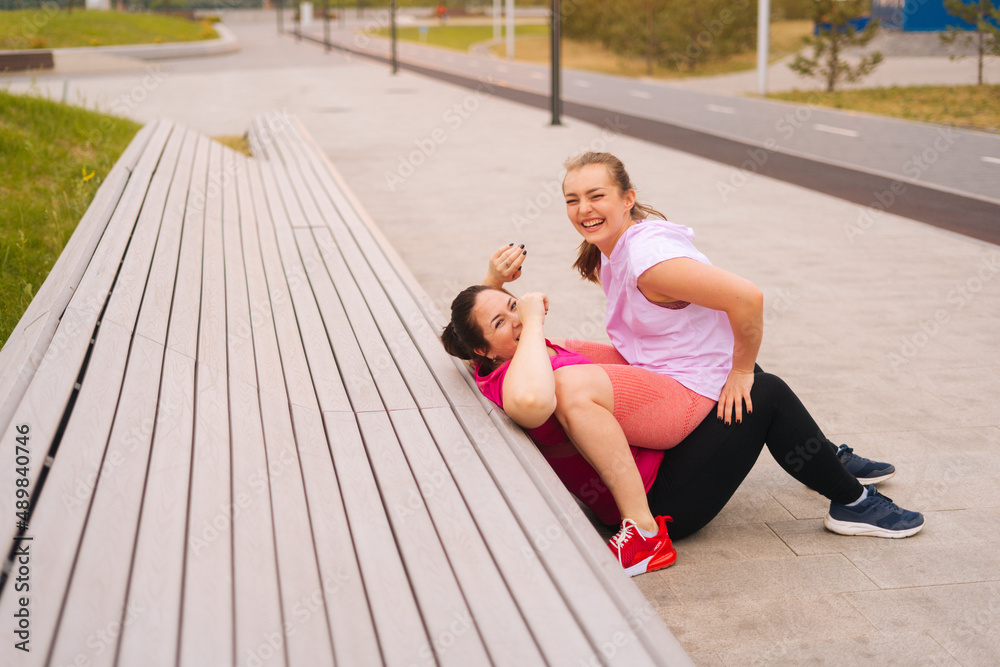 Laughing fat young woman training with cheerful fitness instructor using bench at outdoors gym in city park. Personal training from athletic girl for happy fat young woman with big abdomen outside