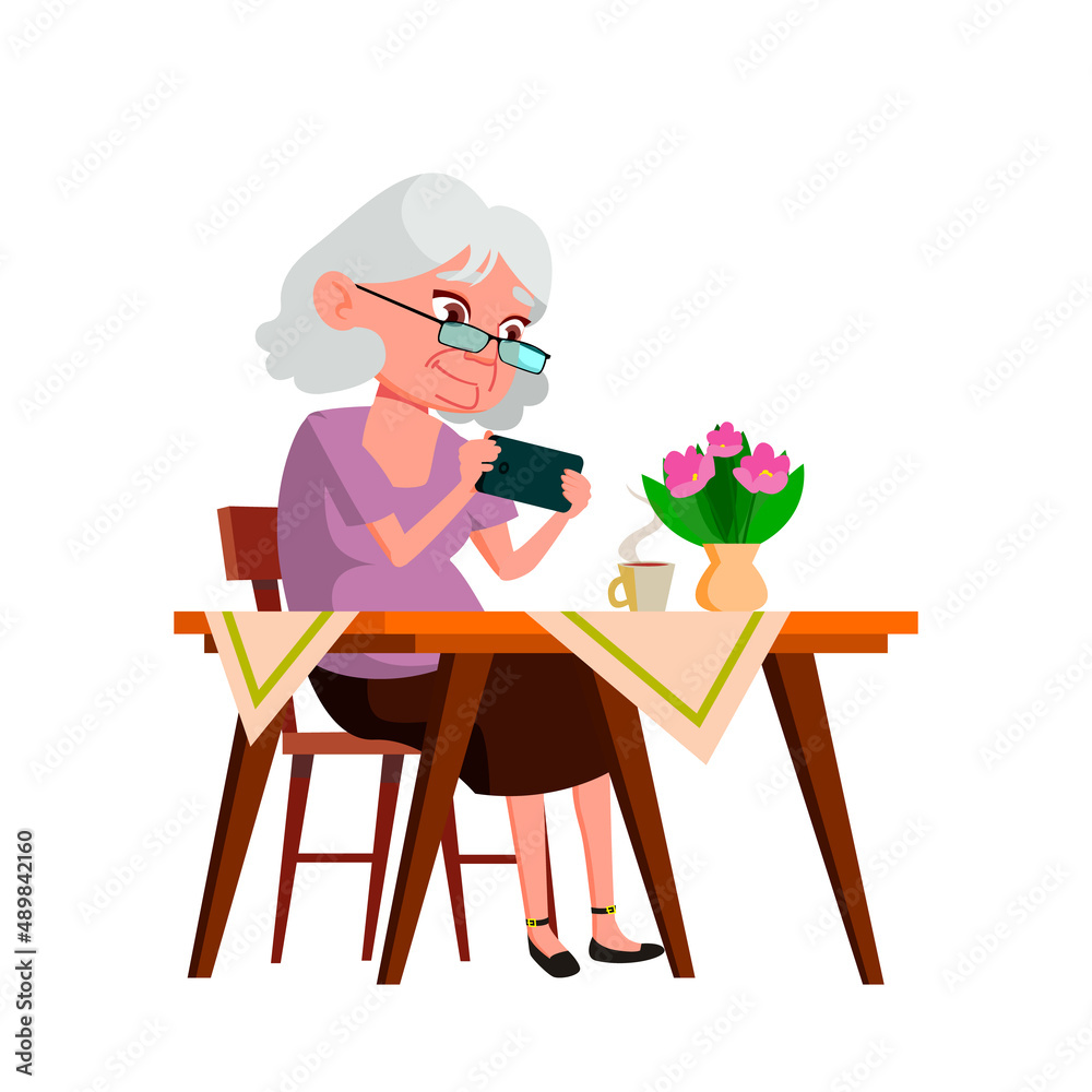 Fototapeta premium Old Woman Grandma Playing On Smartphone Vector. Elderly Lady Sitting At Table, Drinking Hot Drink And Play On Smartphone Digital Device. Character Use Phone At Desk Flat Cartoon Illustration