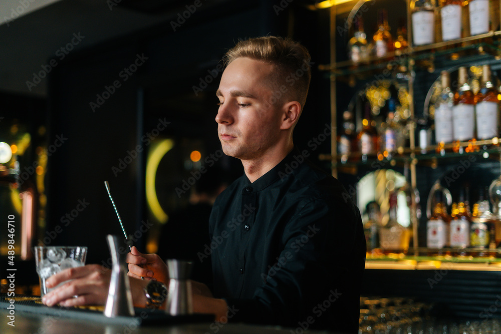 Professional young barman standing behind bar counter and holding stirring spoon and glass filled with ice cubes, preparing to make cocktail, on background of shelves with alcoholic drinks.