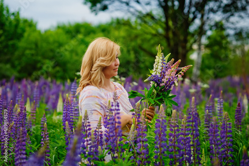 Portrait of a young plump blonde woman in a blooming field of lupines. A woman collects a bouquet of lilac-pink flowers in a meadow.