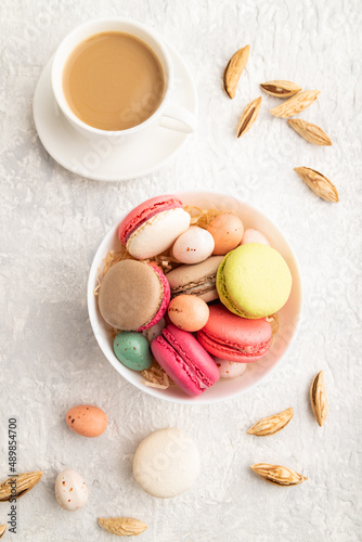 Different colors macaroons and chocolate eggs in ceramic bowl, cup of coffee on gray concrete background. top view, close up.