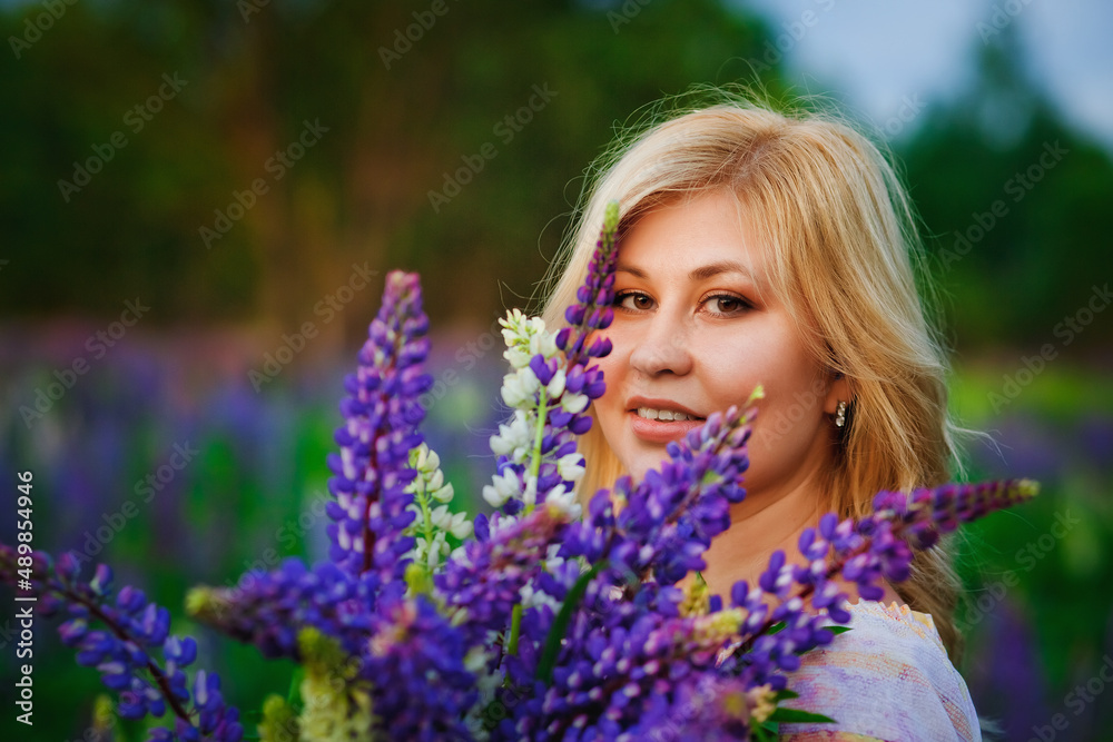 Portrait of a beautiful plus-size blonde with a bouquet of lilac-purple lupine flowers in her hands. A stout woman stands on a blooming meadow. Portrait of a beautiful plus-size blonde with a bouquet of lilac-purple lupine flowers in her hands. A stout woman stands on a blooming meadow.