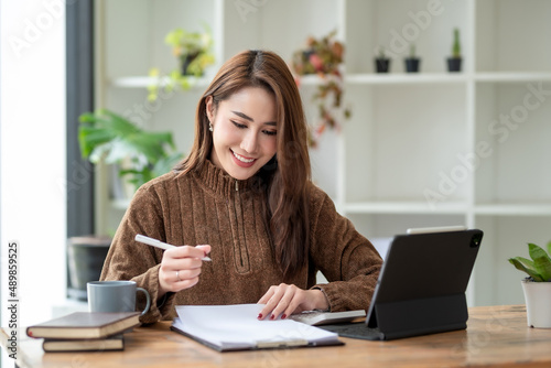 Charming young Asian businesswoman sitting smiling working with document and laptop on the desk in the office.