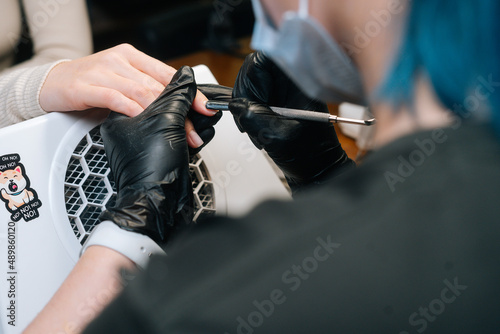 Close-up view from shoulder of unrecognizable manicurist female master removing cuticles by professional metal clippers in salon. Beautician in protective gloves cutting skin on fingernails by nippers