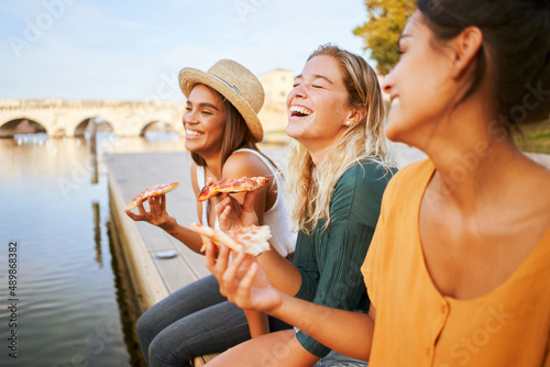 Three beautiful women eating pizza outdoors. The happy girls enjoy the weekend summer vacation together.