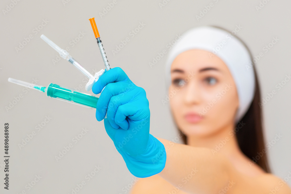 A blurry portrait of a woman with a gloved hand holding three different syringes. Gray background. The concept of beauty and rejuvenation injections A blurry portrait of a woman with a gloved hand holding three different syringes. Gray background. The concept of beauty and rejuvenation injections