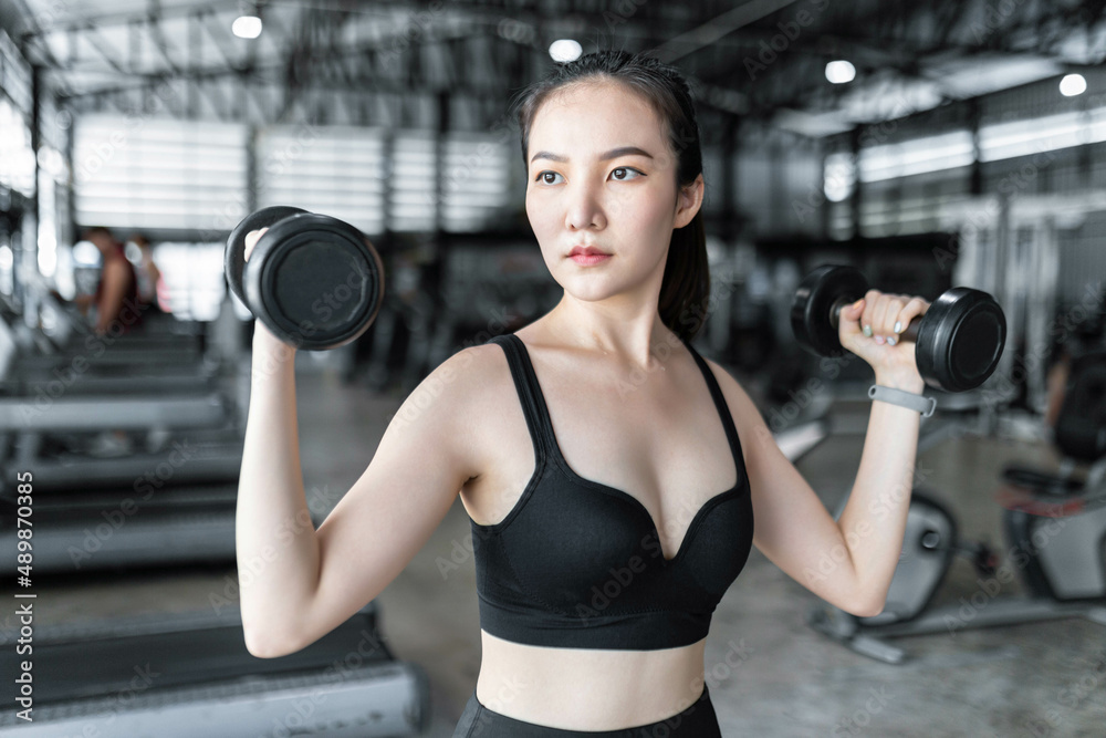 Fototapeta premium exercise concept The healthy woman in dark sport clothes tying her hair up and working out with weightlifting, carrying two black dumbbells up