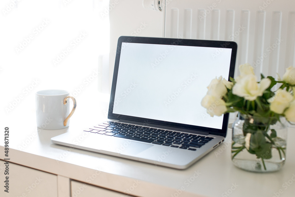 Laptop with white blank screen on white work table. White interior home office concept. Female office desk workspace with white roses and cup of coffee. Close up on screen laptop. Mock up laptop Laptop with white blank screen on white work table. White interior home office concept. Female office desk workspace with white roses and cup of coffee. Close up on screen laptop. Mock up laptop