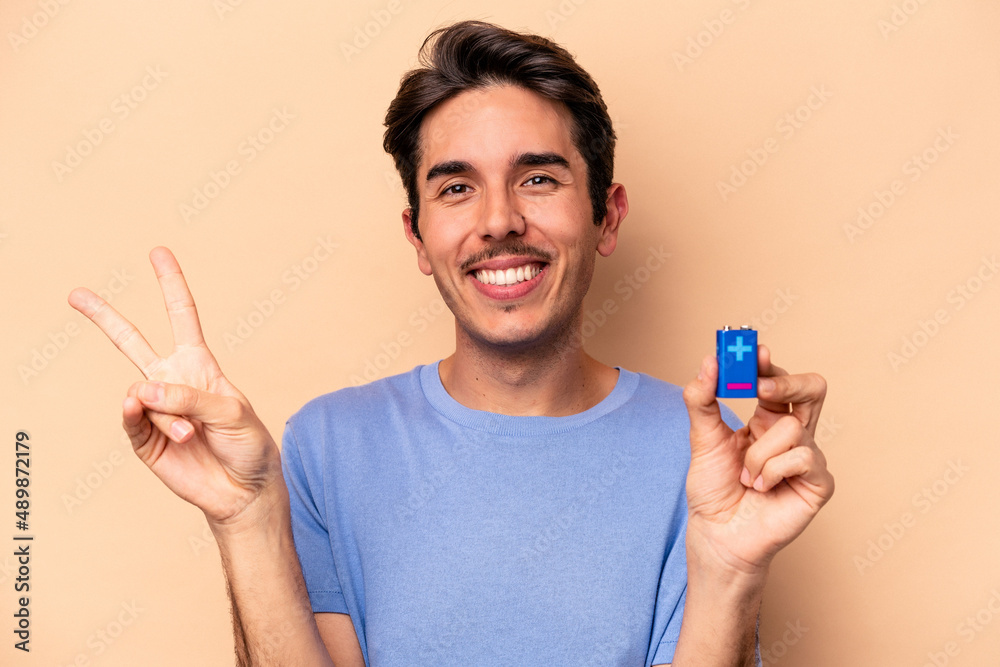 Young caucasian man holding a batterie isolated on beige background joyful and carefree showing a peace symbol with fingers. Young caucasian man holding a batterie isolated on beige background joyful and carefree showing a peace symbol with fingers.