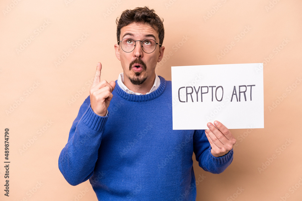 Young caucasian man holding a crypto art placard isolated on beige background pointing upside with opened mouth. Young caucasian man holding a crypto art placard isolated on beige background pointing upside with opened mouth.