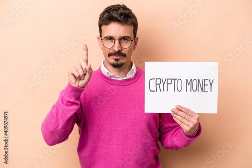 Young caucasian man holding a crypto money placard isolated on beige background showing number one with finger. © Asier