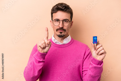 Young caucasian man holding batteries isolated on beige background showing number one with finger.