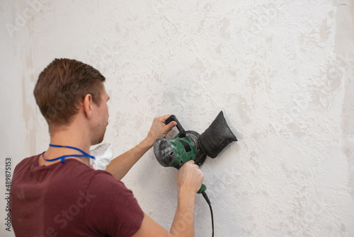 a young man in protective mask and glasses working with sander for smoothing wall surface, polishes surface after putty for painting. Using hand polishing machine for cement wall