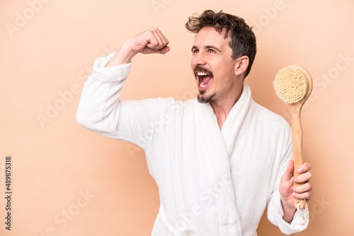 Young caucasian man holding back scratcher isolated on beige background raising fist after a victory, winner concept.