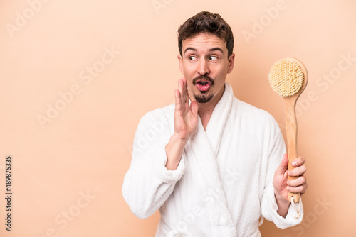 Young caucasian man holding back scratcher isolated on beige background is saying a secret hot braking news and looking aside