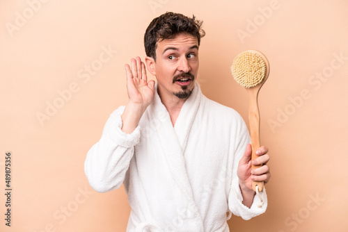 Young caucasian man holding back scratcher isolated on beige background trying to listening a gossip.