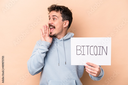 Young caucasian man holding a bitcoin placard isolated on beige background shouting and holding palm near opened mouth.