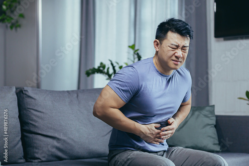 Asian man himself at home sick sitting on the couch, has severe abdominal pain, man in the living room