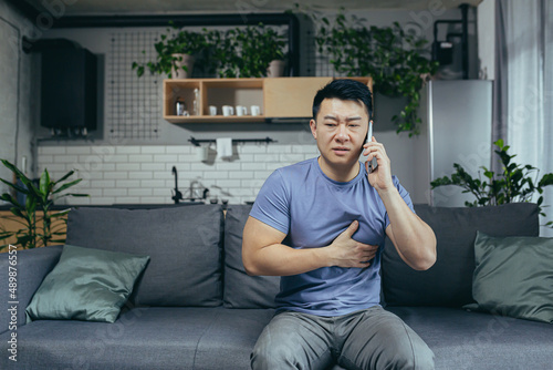 Asian man at home alone, has severe chest pain, calls a doctor, reports his illness, sitting on the couch at home