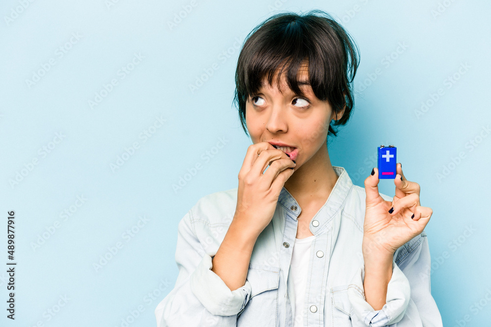 Young hispanic woman holding a batterie isolated on blue background relaxed thinking about something looking at a copy space. Young hispanic woman holding a batterie isolated on blue background relaxed thinking about something looking at a copy space.