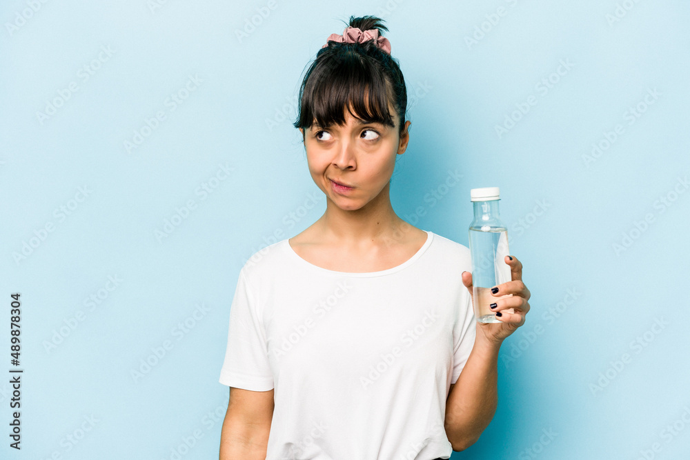 Young hispanic woman holding a bottle of water isolated on blue background confused, feels doubtful and unsure. Young hispanic woman holding a bottle of water isolated on blue background confused, feels doubtful and unsure.