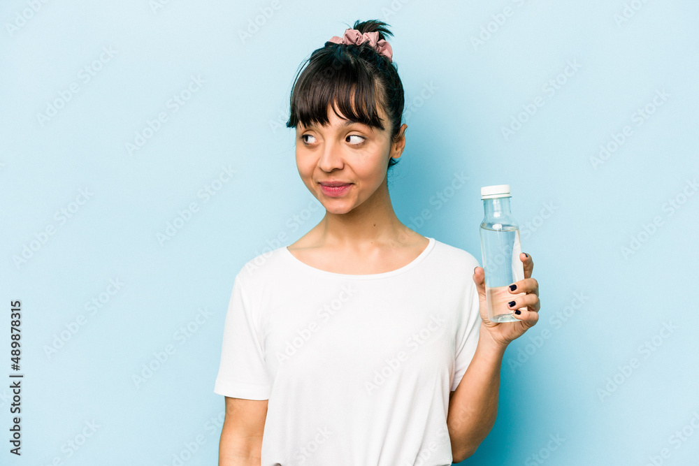Young hispanic woman holding a bottle of water isolated on blue background looks aside smiling, cheerful and pleasant. Young hispanic woman holding a bottle of water isolated on blue background looks aside smiling, cheerful and pleasant.