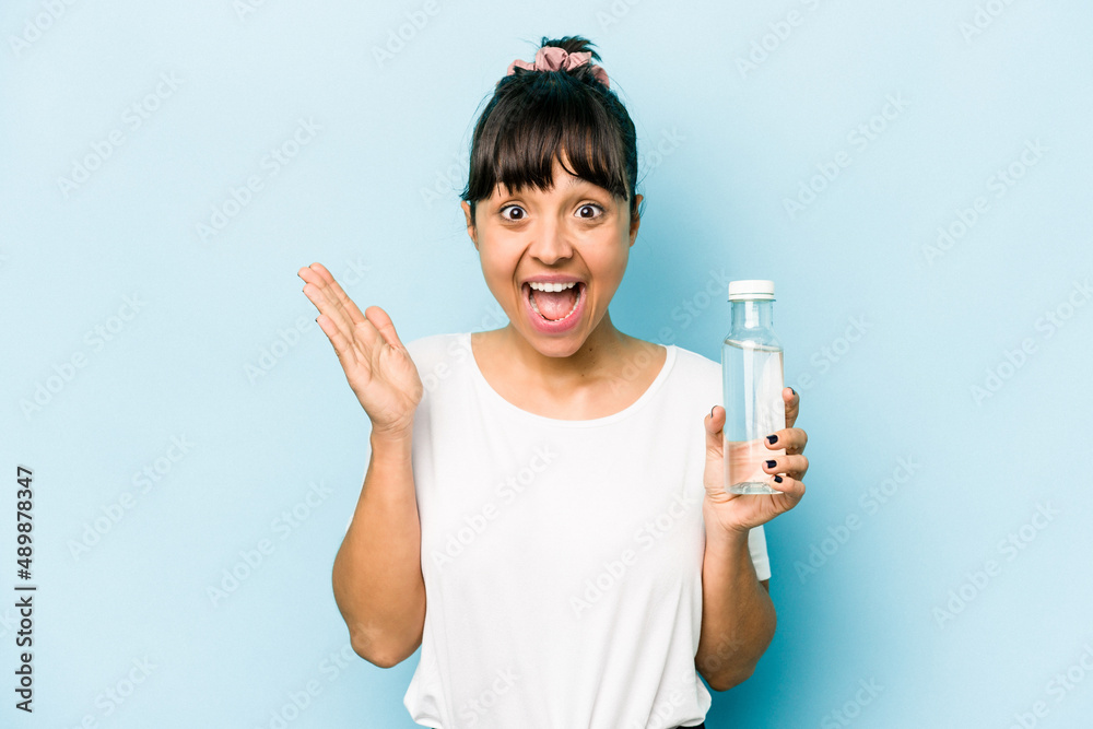Young hispanic woman holding a bottle of water isolated on blue background receiving a pleasant surprise, excited and raising hands. Young hispanic woman holding a bottle of water isolated on blue background receiving a pleasant surprise, excited and raising hands.