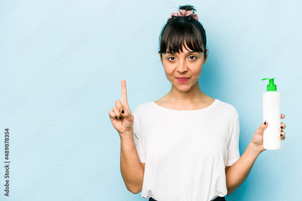 Young hispanic woman holding a body lotion isolated on blue background showing number one with finger. Young hispanic woman holding a body lotion isolated on blue background showing number one with finger.