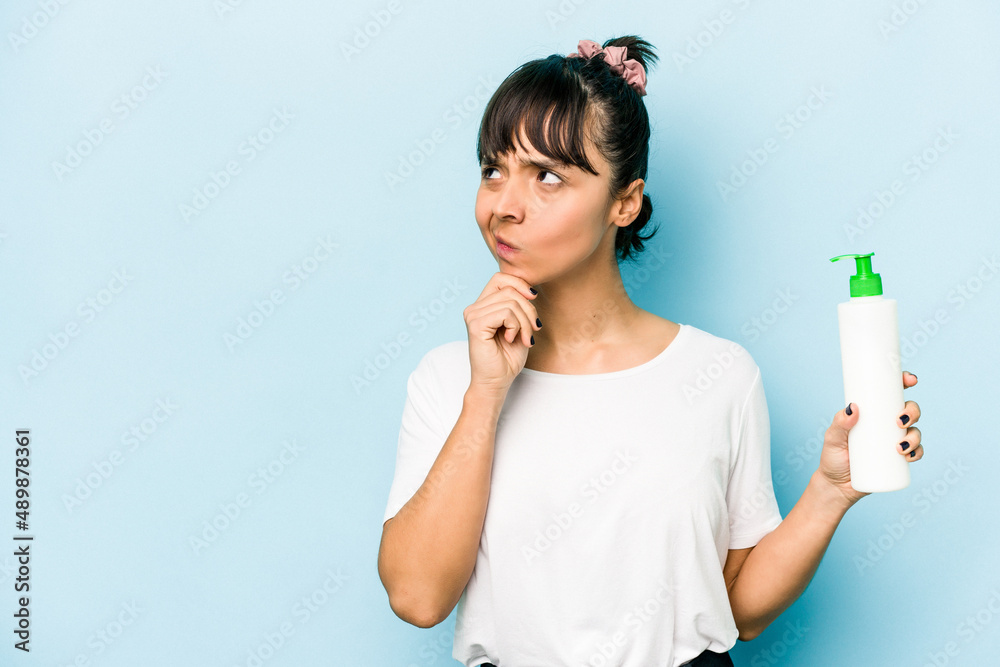 Young hispanic woman holding a body lotion isolated on blue background looking sideways with doubtful and skeptical expression. Young hispanic woman holding a body lotion isolated on blue background looking sideways with doubtful and skeptical expression.