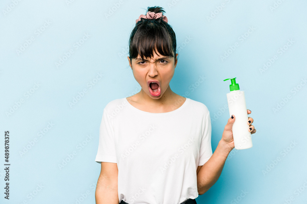 Young hispanic woman holding a body lotion isolated on blue background screaming very angry and aggressive. Young hispanic woman holding a body lotion isolated on blue background screaming very angry and aggressive.