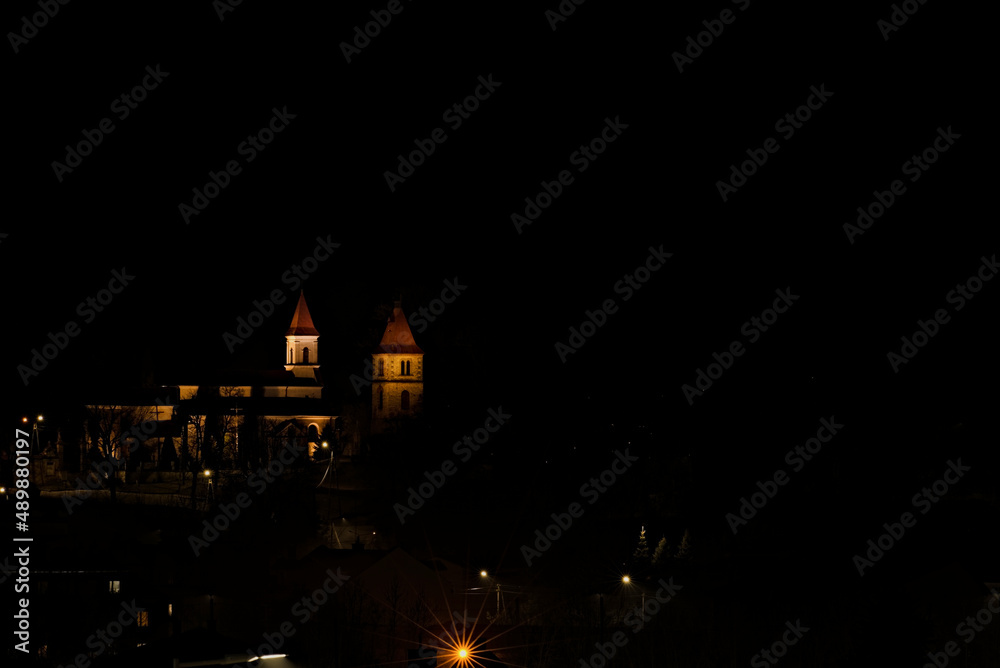 Kościół , pod wezwaniem św. Władysława , w Kunowie ( woj Świętokrzyskie ) ,  nocą , oświetlony , na tle świateł miasta . - obrazy, fototapety, plakaty 